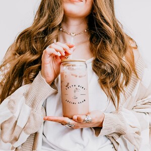 May include: A clear glass tumbler filled with a creamy beverage, featuring the text "SALTY GIRL SOCIAL CLUB." The tumbler has a wooden lid and a straw. The image is of a person holding the glass.