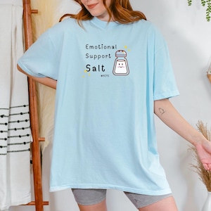 May include: A light blue t-shirt with the text "Emotional Support Salt" and a cartoon salt shaker with a smiley face. The text "#NOTS" is below the salt shaker.