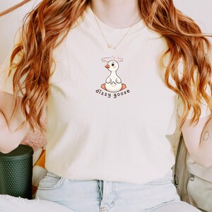 May include: Cream-colored t-shirt featuring a cartoon goose with a halo, sitting cross-legged. The text "dizzy goose" is below the goose. The shirt is being worn by a person with long, wavy auburn hair.