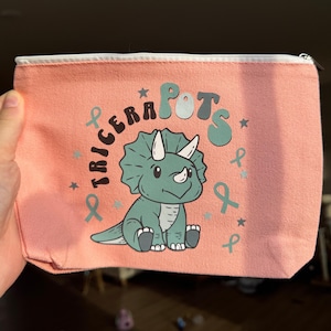 May include: A pink canvas zippered pouch with a white zipper. The pouch features a green triceratops illustration with the text "TRICERAPOTS" in a retro font.