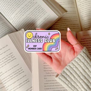 Könnte beinhalten: Ein rechteckiger Aufkleber mit dem Text "Chronic Illness Club VIP Member Card" in Weiß und Lila. Der Aufkleber zeigt ein lächelndes gelbes Gesicht, einen Regenbogen und Sternakzente. Der Aufkleber wird über einem offenen Buch gehalten.
