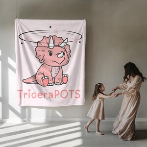 May include: Pink and white fleece blanket with a cartoon pink triceratops with a halo and stars. The text "TriceraPOTS" is printed below the dinosaur.