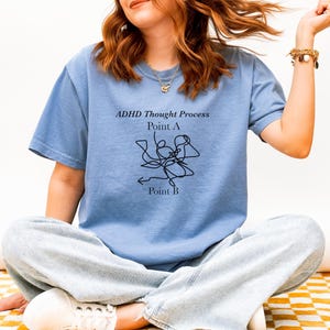 Peut inclure: Un t-shirt bleu clair avec le texte "ADHD Thought Process" et un dessin au trait noir. Le design comprend "Point A" et "Point B" avec une flèche. Le t-shirt est porté avec un jean bleu clair.