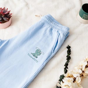 Puede incluir: Pantalones de chándal azul claro con un gráfico de dinosaurio verde y el texto "TriceraPOTS" impreso en la pierna izquierda.