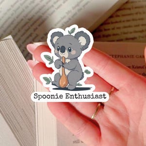 Könnte beinhalten: Aufkleber mit einem Cartoon-Koala, der einen Holzlöffel hält. Der Koala ist grau mit weißem Bauch und grünen Blättern. Der Text "Spoonie Enthusiast" steht darunter.