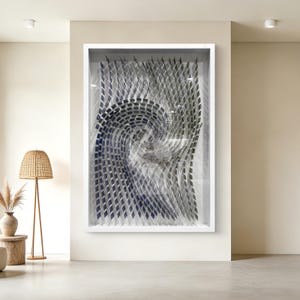 May include: Abstract wall art in a white frame. The artwork features a swirling pattern of layered, geometric shapes in shades of blue, gray, and white, creating a three-dimensional effect. The piece is displayed in a modern, minimalist interior.