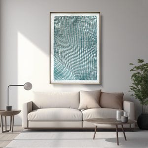 May include: A framed abstract art print with a teal and white grid pattern hangs above a beige sofa. The artwork is in a gold frame. The room is decorated with a modern lamp, a small side table, and a potted plant.