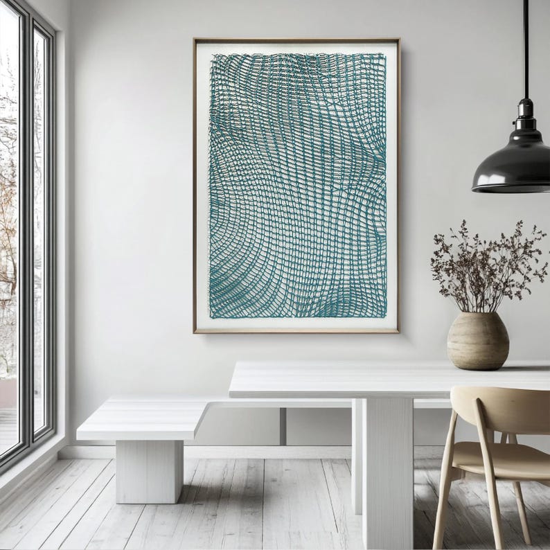 May include: A framed abstract art print featuring a teal-colored, woven-like pattern. The artwork is displayed in a room with white furniture, a wooden floor, and a large window. The print is rectangular and has a thin, brown frame.