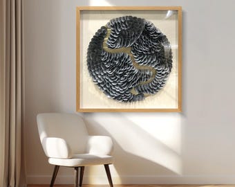 Op de afbeelding: Abstracte wandkunst in een natuurlijke houten lijst. Het kunstwerk heeft een cirkelvormig ontwerp dat is samengesteld uit gelaagde grijstinten met een goudkleurig accent. Het stuk wordt gepresenteerd boven een witte fauteuil met donkere houten poten.
