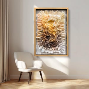 Puede incluir: Obra de arte abstracta enmarcada en un marco de madera. La obra presenta papel en capas en tonos beige, marrón y dorado, creando un efecto texturizado y tridimensional. La pieza rectangular cuelga de una pared blanca, con una silla blanca en primer plano.