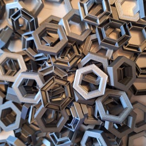 Puede incluir: Una colección de formas hexagonales geométricas grises. Las formas están impresas en 3D y tienen un diseño en capas. Los objetos están dispersos y se superponen, creando una superficie texturizada. La paleta de colores es monocromática, con variaciones de gris.