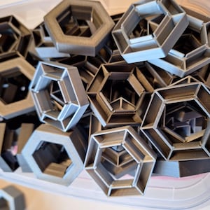 Può includere: Una collezione di oggetti esagonali geometrici grigi con un design aperto e multistrato. Gli oggetti sono impilati all'interno di un contenitore di plastica trasparente, suggerendo un contesto di produzione o stoccaggio. Le forme hanno un aspetto metallico.