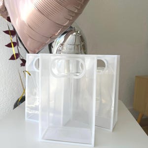 May include: Clear rectangular gift bags with white trim and handles are arranged on a white surface. Two metallic balloons in rose gold and silver are in the background. The bags are empty and ready to be filled.