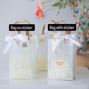 May include: Two clear plastic favor bags with white tissue paper inside. The bag on the left has a wooden handle and a white ribbon with a "Thank You" tag. The bag on the right has a wooden handle, a white ribbon, and a round sticker that says "Eliza's Baby Shower".