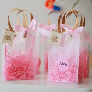 May include: Clear plastic favor bags with pink tissue paper, a pink ribbon, and a gold "Thank You" tag. One bag has a pink circle sticker with the name "Abby" on it.