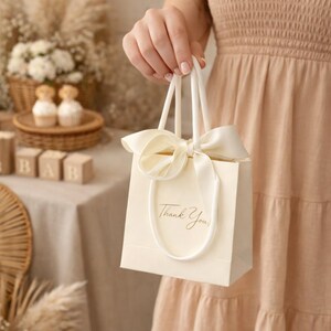 Puede incluir: Bolsa de regalo marfil con lazo y asas color crema. La bolsa tiene las palabras "Thank You" en escritura elegante. La bolsa se sostiene frente a una persona que lleva un vestido color melocot&oacute;n.