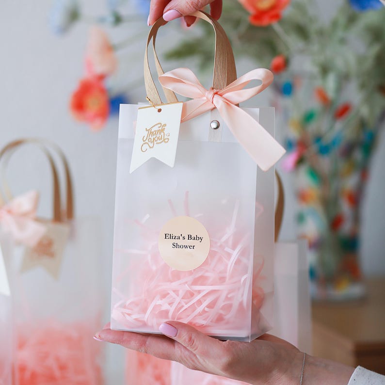 May include: A clear gift bag with a peach ribbon and handles. The bag contains pink shredded paper and a round sticker that reads "Eliza's Baby Shower." A small tag with "Thank You" is attached.