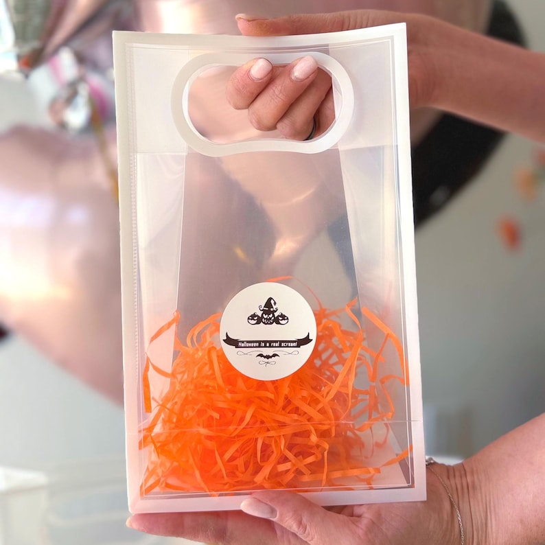 Peut inclure: Un sac cadeau en plastique transparent avec une poign&eacute;e, rempli de papier d&eacute;chiquet&eacute; orange. Un autocollant rond avec un motif d'Halloween et le texte "Halloween is a real scream!" est appos&eacute; sur le sac.