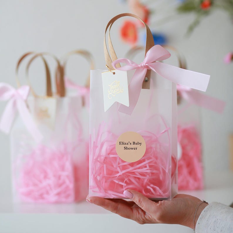 May include: Clear plastic favor bags with pink tissue paper and a pink ribbon. The bags have a gold sticker that says "Eliza's Baby Shower".