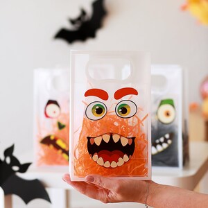 May include: Clear plastic Halloween treat bags with monster faces. The bags have orange, green, and black monster designs. The bags are filled with orange paper shred. A black bat decoration is in the background.