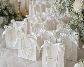Op de afbeelding: Een verzameling kleine witte cadeauzakjes met witte touwgrepen en een witte satijnen strik aan de bovenkant. Elk zakje is aan de voorzijde bedrukt met de gouden cursieve tekst Thank You en staat in rijen op een gedrapeerde witte stoffen ondergrond.