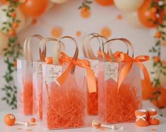 Könnte beinhalten: Transparente Geschenktüten gefüllt mit orangefarbenem Papier, gebunden mit orangefarbenen Bändern und Anhängern mit der Aufschrift "Thank you". Die Tüten haben braune Papiergriffe. Der Hintergrund umfasst orangefarbene und cremefarbene Ballons und orangefarbene Dekorationen.