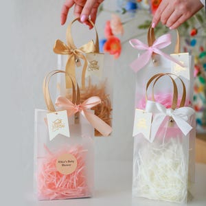 May include: Five clear plastic gift bags with handles, filled with pink and white tissue paper. Each bag has a gold ribbon and a small tag with gold lettering. The tags say "Thank You" and "Eliza's Baby Shower".