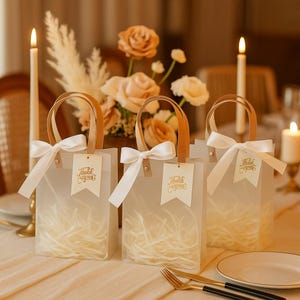 May include: Three translucent gift bags with white bows and tan leather handles are arranged on a table. Each bag has a tag that reads "Thank you" in gold script. The bags are filled with shredded paper.