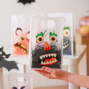 May include: Clear plastic Halloween treat bags with monster faces. The bags have a handle and are filled with black paper shred. The monster faces are orange, green, and white.