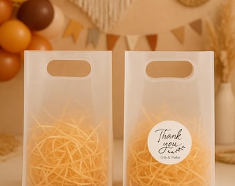May include: Two transparent gift bags filled with yellow shredded paper. One bag has a white sticker with the words "Thank you" and the names "Gla & Pieter". The bags have a handle cut-out.