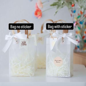May include: Clear gift bags with white ribbon bows and paper handles. One bag has a "Thank You" tag, another has a sticker that says "Eliza's Baby Shower". The bags are filled with shredded paper.
