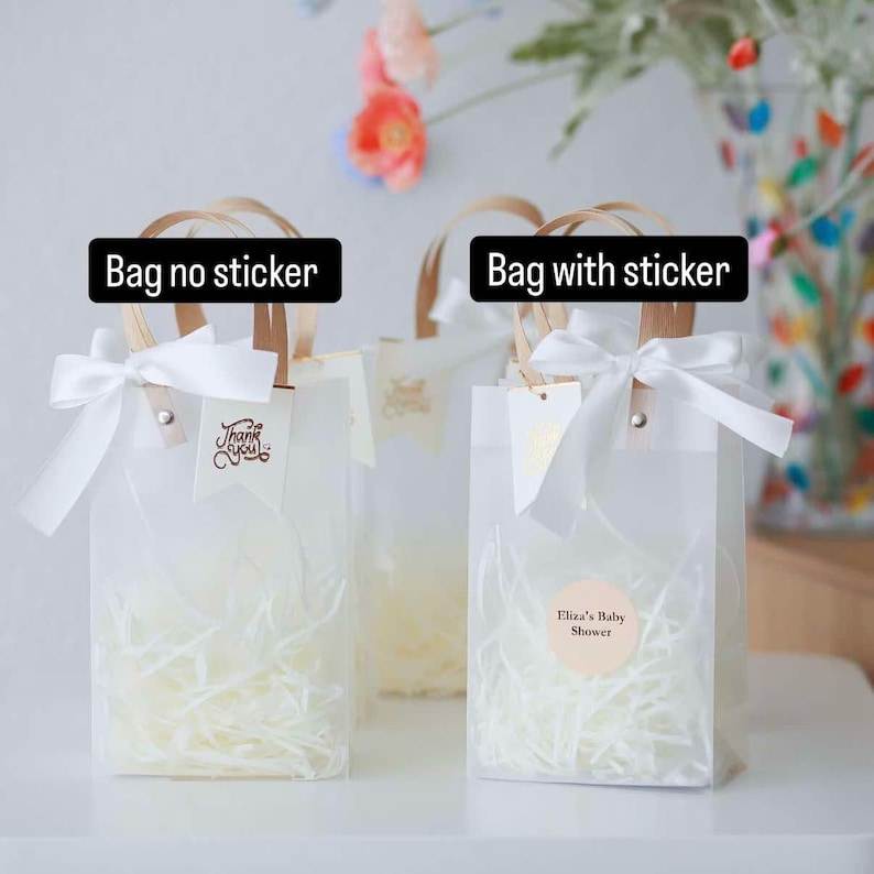 May include: Clear gift bags with white ribbon bows and paper handles. One bag has a "Thank You" tag, another has a sticker that says "Eliza's Baby Shower". The bags are filled with shredded paper.