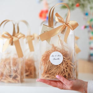 May include: Clear plastic gift bag with a gold ribbon and a white circle label that says "Mr. & Mrs. Smith". The bag is filled with shredded paper.