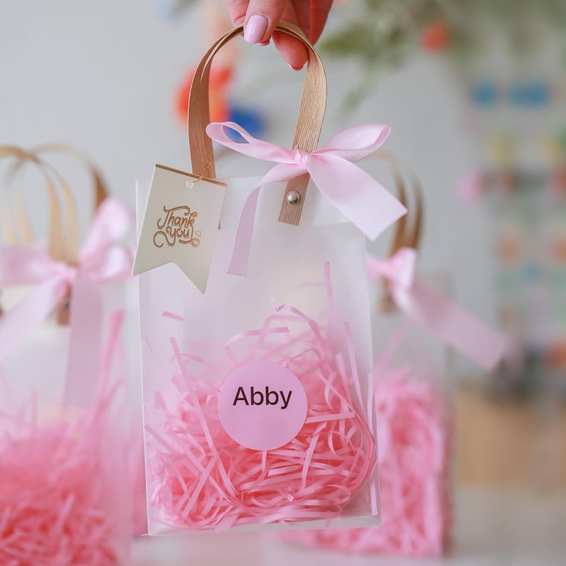 May include: Pink and white favor bags with gold lettering. The bags have a clear window and are filled with pink tissue paper. One bag has a tag that says "Thank You" and another bag has a tag that says "Abby".