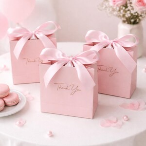 Puede incluir: Tres bolsas de regalo rosas con lazos de sat&eacute;n, cada una con la inscripci&oacute;n dorada "Thank You". Las bolsas est&aacute;n sobre una superficie blanca, con macarons rosas y un jarr&oacute;n de rosas rosas en el fondo. Confeti y perlas rosas esparcidas.