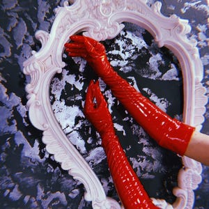 May include: A pair of long, shiny red gloves are positioned within a white, ornate heart-shaped frame. The frame is against a textured background of black, silver, and gray. The gloves are the focal point, with their glossy surface reflecting light.