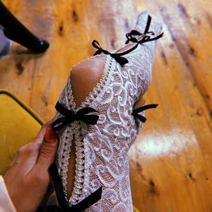 May include: White lace stockings featuring a vertical cutout design along the front. The edges of the cutout are trimmed with decorative scalloped lace and secured with four small black ribbon bows placed at intervals along the length.