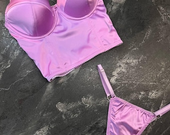 May include: A lilac satin lingerie set, comprising a bustier top and a thong. The bustier has structured cups and adjustable straps. The thong features thin straps and a delicate design. The set is made of a smooth, shiny fabric, in a soft lilac colour.