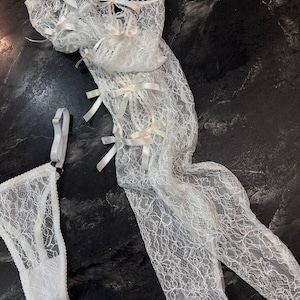 May include: White lace lingerie set featuring a garter belt, thigh-high stockings, and a thong. The delicate lace is adorned with small satin bows. The set is displayed on a dark, textured surface.