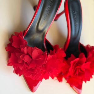 May include: A pair of red high-heeled mules featuring a pointed toe and a glossy finish. The front of each shoe is adorned with a cluster of layered red fabric flower petals. The black insole is printed with the word Fashion in script.