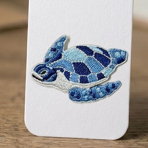 May include: Embroidered sea turtle patch in shades of blue and white. The turtle has a patchwork design on its shell and flippers. The patch is on a white background.