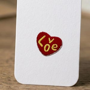 May include: A white rectangular card featuring a small, embroidered red heart patch centered on its surface. The heart contains the yellow embroidered text "Loe" with the letter "v" stylized as a small checkmark shape within the heart design.
