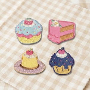 Peut inclure: Quatre écussons brodés représentant des desserts. On y trouve un cupcake avec un glaçage bleu et des vermicelles, une part de gâteau rose avec une fleur, un dessert à la crème anglaise avec une cerise et un cupcake avec un glaçage bleu et une cerise.