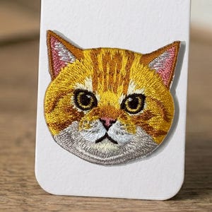 May include: Embroidered patch featuring a detailed cat face. The cat has orange and yellow fur with brown stripes, large black eyes, and a pink nose and ears. The patch is likely for clothing or accessories.