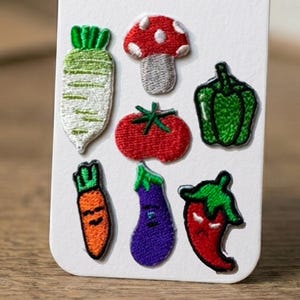 May include: A set of seven embroidered patches featuring a white radish with green leaves, a red and white mushroom, a green bell pepper, a red tomato, an orange carrot with a face, a purple eggplant with a face, and a red chili pepper with a face.