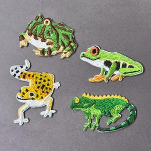 May include: Four embroidered patches featuring amphibians and reptiles. The patches include a green and brown frog, a green frog with red eyes, a yellow and white gecko, and a green iguana with a yellow crest.
