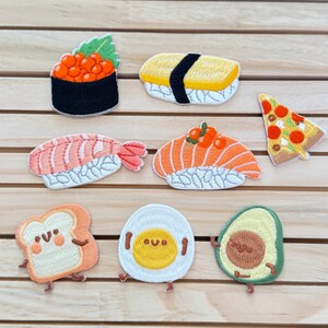 Può includere: Nove toppe ricamate da stirare che rappresentano diversi cibi, tra cui sushi, pizza, avocado, uova e pane. Le toppe sono su uno sfondo di legno.