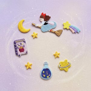 May include: Embroidered patches featuring a witch on a broom, a crescent moon, stars, a potion bottle, and a spell book. The witch wears a blue dress and red bow. The patches are in shades of yellow, blue, purple, and brown.