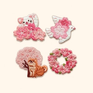 May include: Four embroidered patches featuring pink cherry blossoms. One shows a white cat, one a white bird, one a brown dog sitting by a tree, and one a circular floral wreath. Each patch is detailed with intricate thread work and vibrant colors.