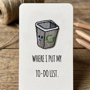 May include: A white card with rounded corners featuring an embroidered grey bin with a green recycling symbol. The text "WHERE I PUT MY TO-DO LIST." is printed below the image. The card is on a wooden surface.